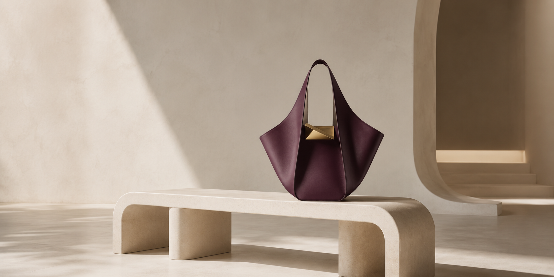 A plum leather tote with a gold clasp on a travertine bench in a sunlit plaster interior